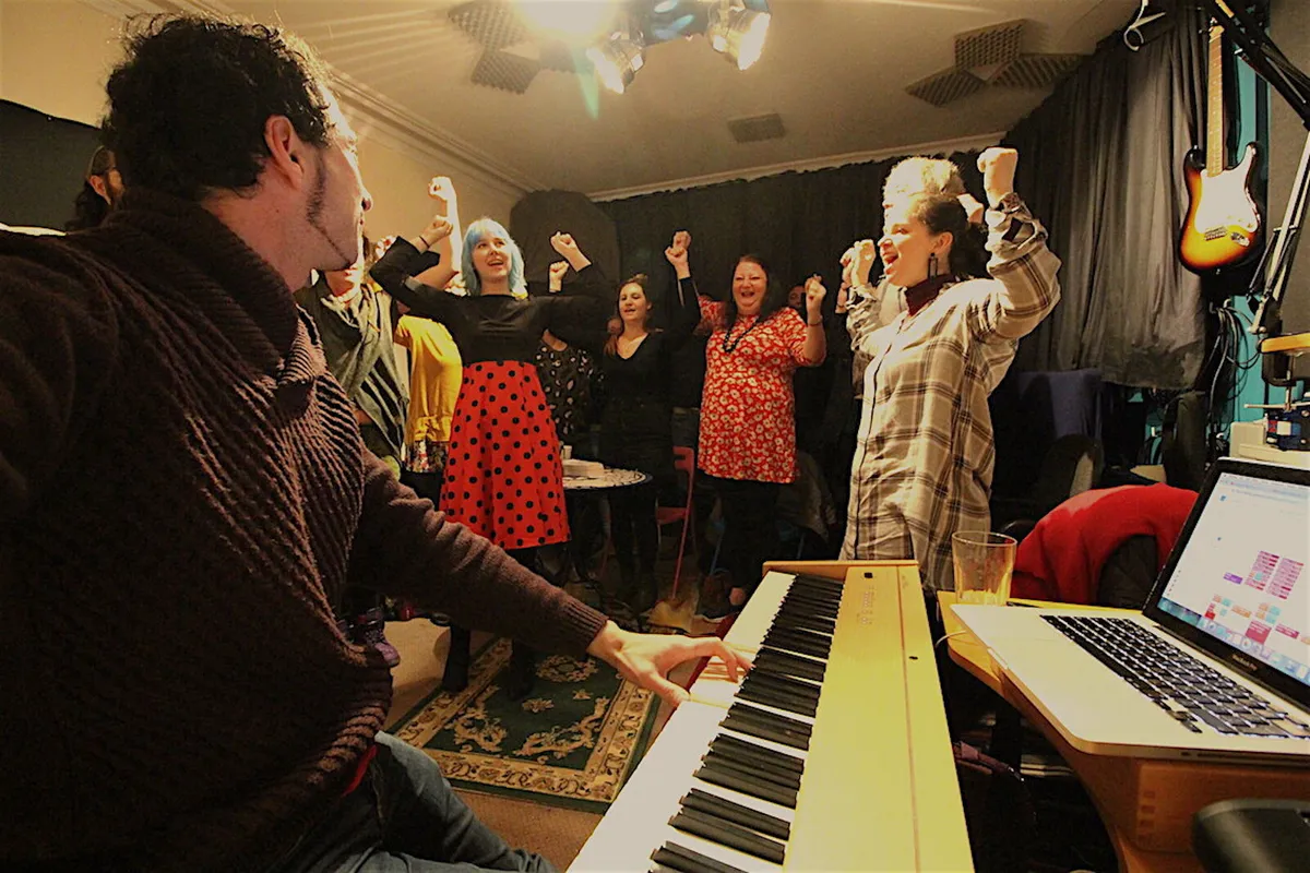 A group of singers with raised fists singing together around a piano at a Discover & Design Your Authentic Voice workshop in Bristol
